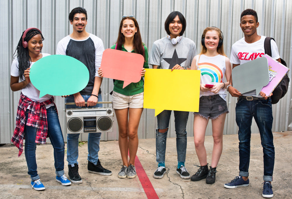 A diverse group of young people standing outdoors, holding colorful speech bubbles and a retro boombox, symbolizing communication and self-expression.