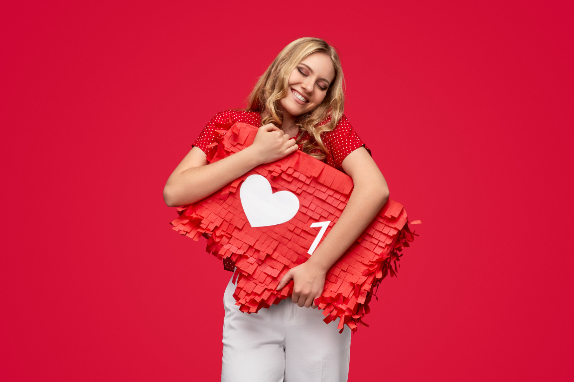 Young woman in red hugging a giant red piñata shaped like a social media notification with a heart and the number one, standing against a solid red background, symbolizing love for social media engagement.