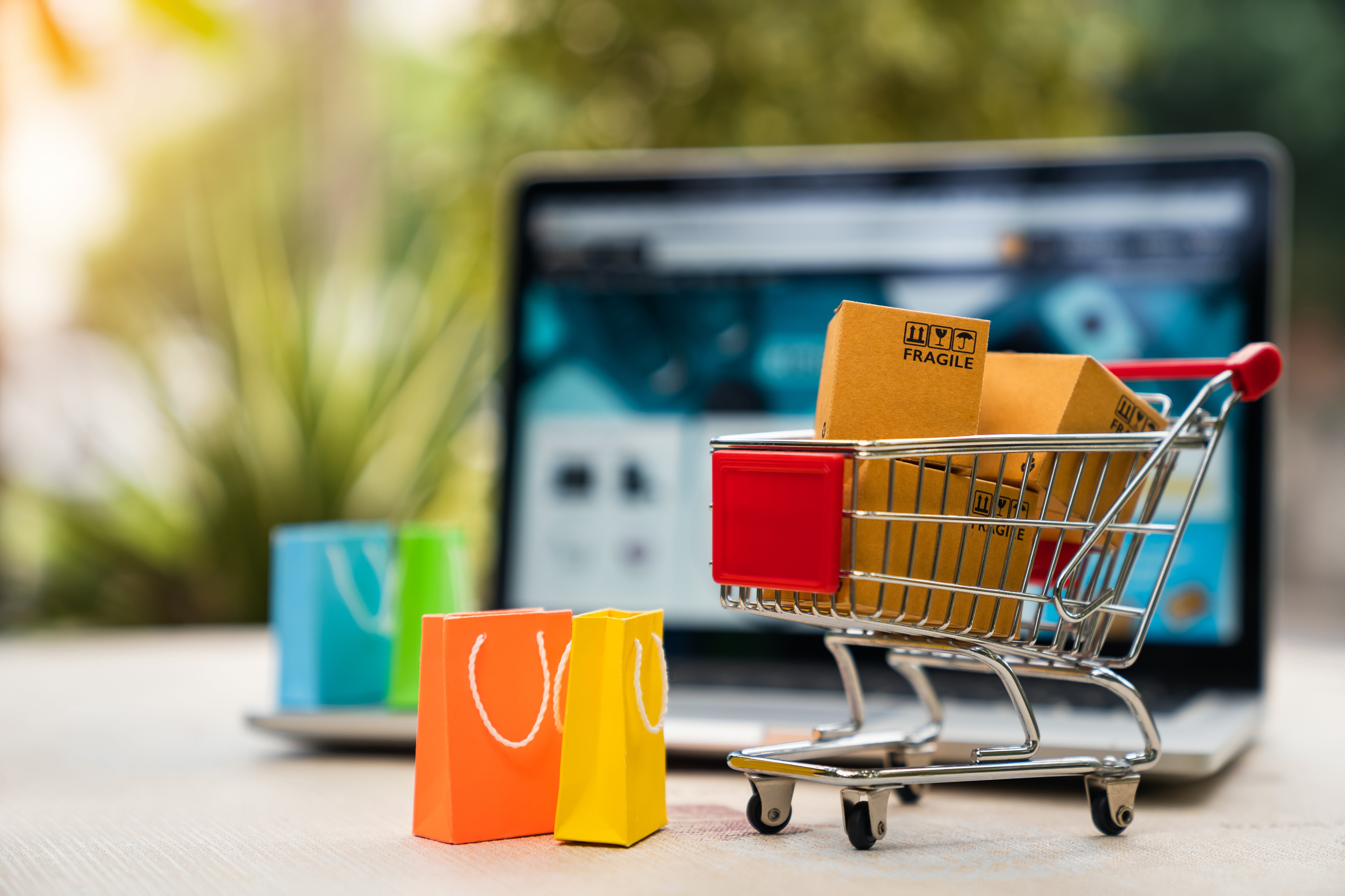 Mini shopping cart filled with packages placed in front of a laptop displaying an online store, symbolizing e-commerce growth and digital shopping trends.